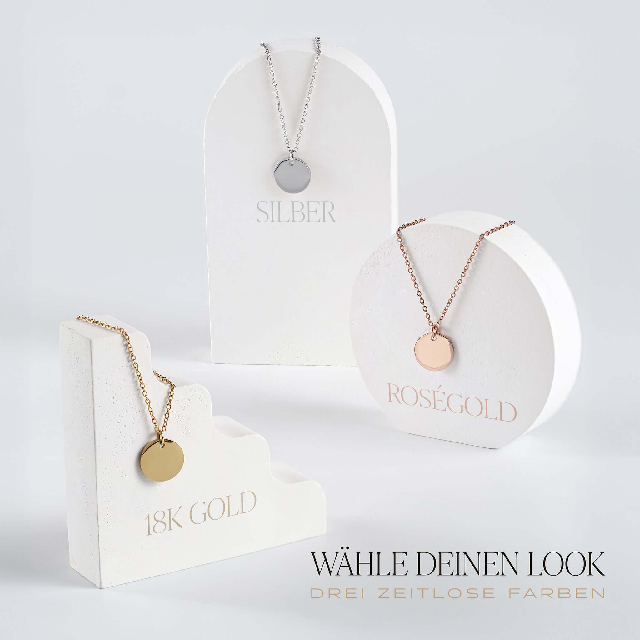 Drei runde Namens-Halsketten in Silber, Roségold und 18K Gold auf weißen Ständern.